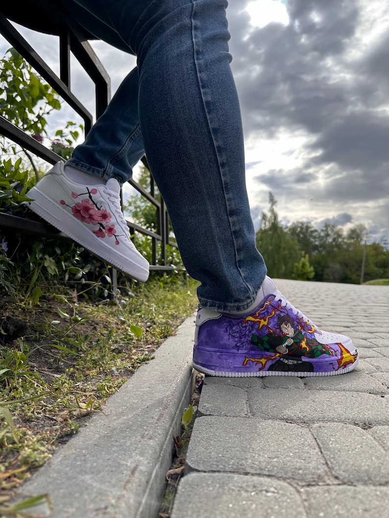 May include: White sneakers with custom artwork. One shoe features pink cherry blossoms, the other a purple and green anime-style design. The shoes are worn with blue jeans.