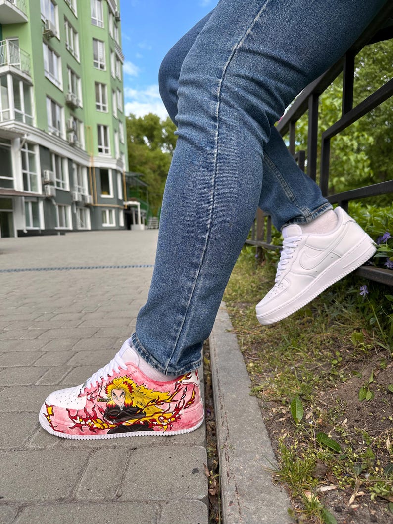 May include: White sneakers with custom artwork. One shoe features a pink and red design with a character illustration and yellow hair. The other shoe is plain white. The person is wearing blue jeans.