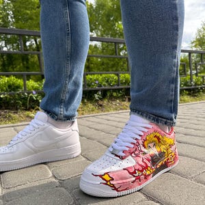 May include: White leather sneakers with a custom design on one shoe. The design features a character with yellow hair and a pink and yellow flame pattern. The other shoe is a plain white sneaker.