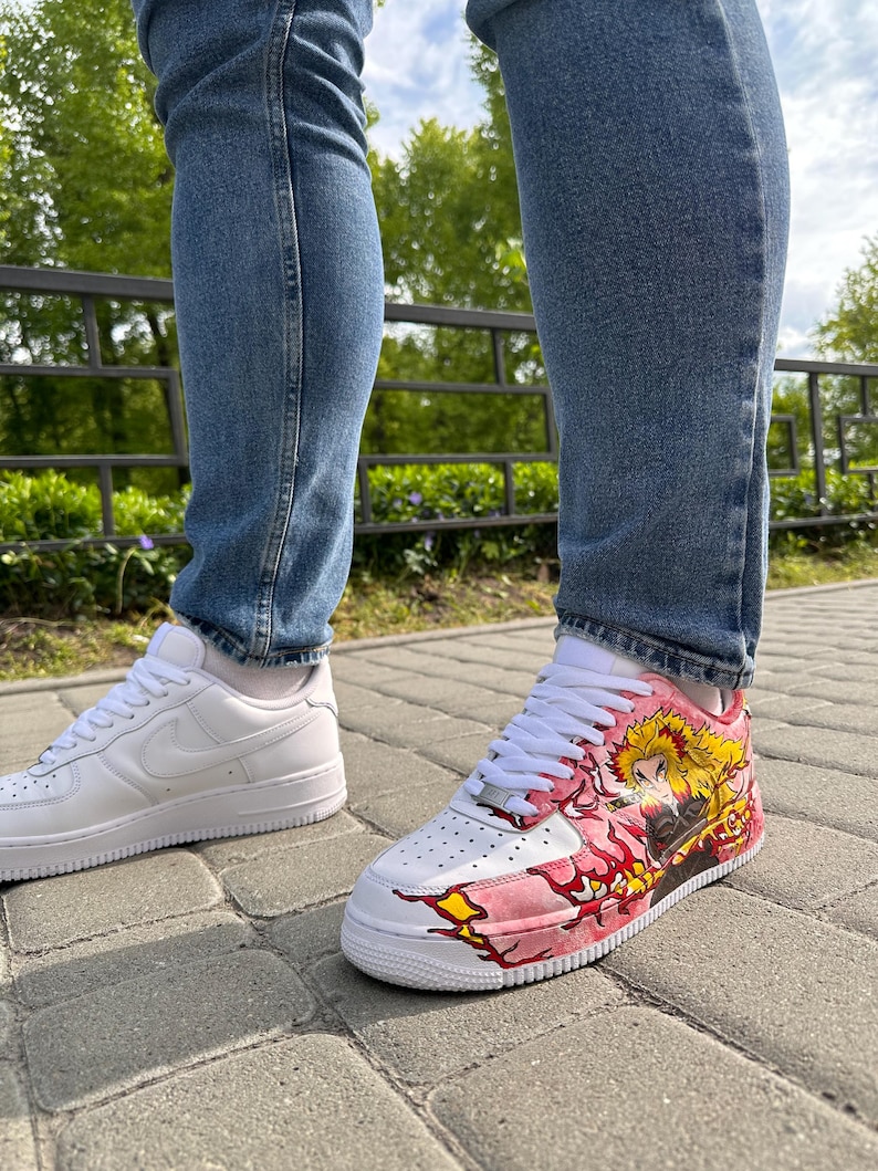 May include: White leather sneakers with a custom design on one shoe. The design features a character with yellow hair and a pink and yellow flame pattern. The other shoe is a plain white sneaker.
