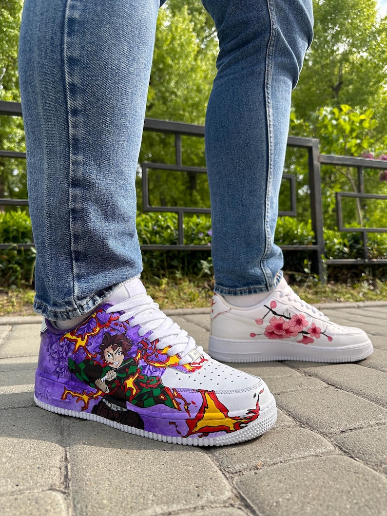 May include: White sneakers with custom artwork. One shoe features a colorful anime character design with purple, green, and yellow accents. The other shoe has pink cherry blossom branch art. The shoes are worn with blue jeans.