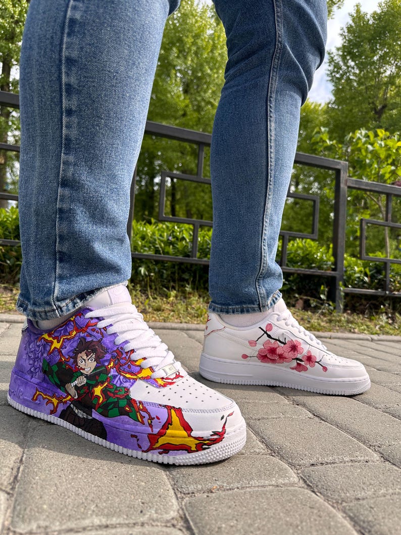 May include: White sneakers with custom artwork. One shoe features a colorful anime character design with purple, green, and yellow accents. The other shoe has a pink cherry blossom design. Both shoes have white laces and soles.