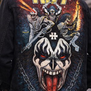 May include: Black denim jacket featuring a colorful graphic of the band KISS. The design includes the band's logo, band members in makeup, and a large face graphic. The jacket has a classic collar and button closure.