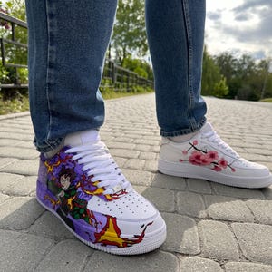 May include: White sneakers with custom artwork. One shoe features a character design with purple, red, and yellow accents. The other shoe has pink cherry blossom branch detailing. Both shoes have white laces and soles. The person is wearing blue jeans.