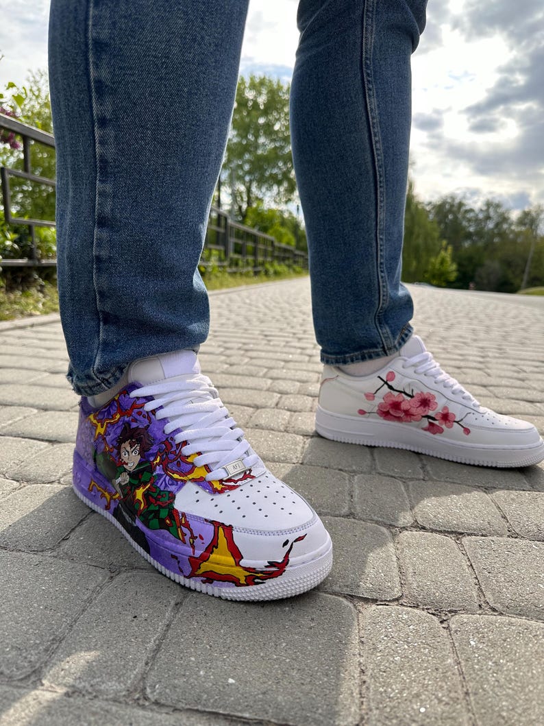 May include: White sneakers with custom artwork. One shoe features a character design with purple, red, and yellow accents. The other shoe has pink cherry blossom branch detailing. Both shoes have white laces and soles. The person is wearing blue jeans.