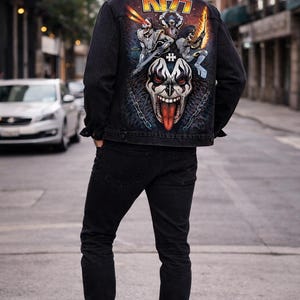 May include: Black denim jacket with a colorful graphic on the back featuring the band KISS. The graphic includes the band's logo and illustrations of the band members. The jacket is paired with black jeans and boots.