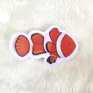 Puede incluir: Una pegatina con un diseño de pez payaso de dibujos animados. El pez tiene rayas naranjas y blancas, con detalles negros en las aletas. La pegatina tiene un borde blanco y está sobre un fondo blanco texturizado.