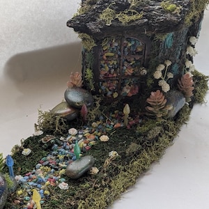 May include: A miniature fairy house with a mossy roof and a window made of colorful glass beads. The house is set on a bed of green moss with a path of colorful pebbles leading to it.