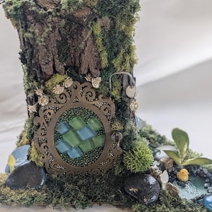 May include: A miniature fairy house made from a tree trunk with a moss covered roof and a decorative door with a mosaic design. The house is surrounded by moss, rocks, and a small pond with a yellow duck.