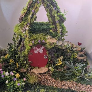 May include: A miniature fairy house with a red door featuring a butterfly design. The house is covered in green moss and bark, with small flowers and a stone pathway leading to the door. The roof has an opening.