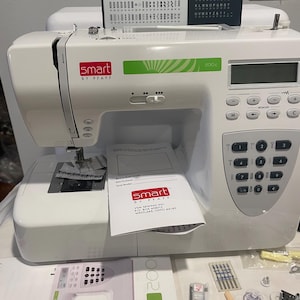 Peut inclure: Machine à coudre blanche Smart by Pfaff 200c avec écran numérique et boutons de commande. La machine a un accent vert et comprend un manuel et des accessoires. Un tableau de motifs et de lettres est visible.