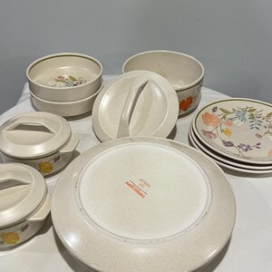 Op de afbeelding: Een verzameling vintage beige serviesgoed met bloemmotieven. De set bevat borden, kommen en serveerschalen met deksels. De borden en kommen hebben een bloemmotief in tinten oranje, geel en paars. De serveerschalen hebben handvatten.