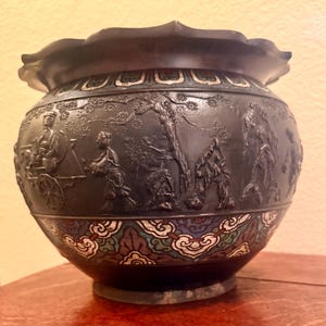 May include: A dark bronze-colored decorative vase with a scalloped rim. The vase features a detailed relief of a landscape with figures and a decorative band with colorful cloud-like patterns.