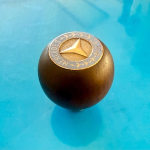May include: A dark brown, spherical Mercedes-Benz gear shift knob. The top features a silver ring with the Mercedes-Benz logo, a gold three-pointed star within a circle, and the word "MERCEDES" in a circular pattern.