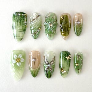 Op de afbeelding: Een verzameling van tien amandelvormige kunstnagels met ingewikkelde ontwerpen. De nagels hebben tinten groen, wit en goud, met bloemen-, vlinder- en abstracte patronen. Sommige nagels zijn versierd met strass-steentjes en metalen accenten, wat een artistieke manicure creëert.