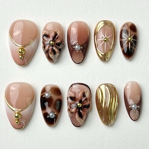 May include: A set of ten almond-shaped false nails. The nails feature a variety of designs, including gold accents, floral embellishments, and pearl accents. The color palette includes shades of brown, gold, and nude.