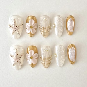 Könnte beinhalten: Ein Set aus zehn künstlichen Nägeln mit Strand-Thema. Designs umfassen Seesterne, Blumen und Muschelmuster in Weiß, Gold und Perlfarben. Einige Nägel weisen 3D-Verzierungen auf.
