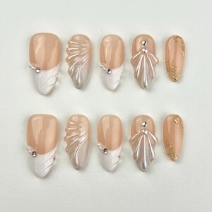 May include: A set of ten artificial nails in various designs. The nails feature a nude base with white tips, pearl accents, and gold leaf details. Some nails have a sculpted, shell-like design. The nails are oval-shaped.