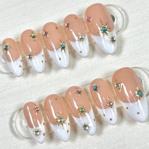 Puede incluir: Diez uñas postizas de color nude con puntas blancas. Cada uña tiene un diseño diferente de pedrería dorada y turquesa.