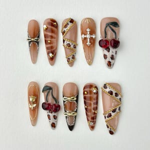 May include: A collection of ten almond-shaped false nails with diverse designs. The nails showcase brown, tan, and leopard print patterns, embellished with gold accents, bows, cherries, and a cross. Some nails have a glossy finish, others a matte appearance.