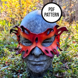 Puede incluir: Una máscara de cuero roja y naranja con un diseño intrincado, usada por un busto de piedra. La máscara tiene una apariencia ardiente, con detalles recortados y un borde rojo oscuro. El busto está situado en un entorno natural al aire libre. El texto "PDF PATTERN" está en un círculo blanco.