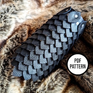 Può includere: Braccialetto in pelle nera con un motivo a scaglie. Il braccialetto è realizzato con singole scaglie di pelle cucite insieme. Il braccialetto ha un'etichetta con la scritta "PDF pattern".