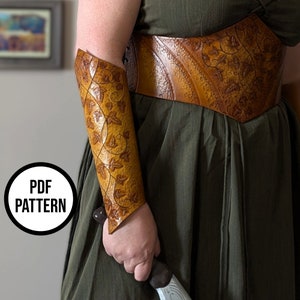 May include: A brown leather arm guard with a vine and leaf pattern. The arm guard is worn on the left arm of a person wearing a green dress and a brown leather belt with a similar vine and leaf pattern. The text "PDF PATTERN" is visible in a circle.