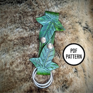 May include: A green leather leaf-shaped accessory with silver metal hardware. The accessory features two leaves with detailed veining, two silver rivets, and a double ring at the bottom. The text "PDF PATTERN" is in a white circle.