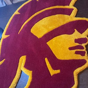 May include: A maroon and gold rug with a stylized image of a Spartan helmet.