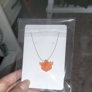 Puede incluir: Un collar de cadena plateada con un colgante de corazón naranja. El colgante tiene una superficie texturizada y parece estar hecho de plástico.