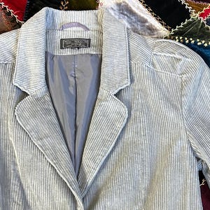 Puede incluir: Blazer de pana gris claro con solapas de muesca, dos bolsillos delanteros y tres botones en cada puño. El blazer tiene cierre de dos botones y forro gris. La tela tiene una textura acanalada vertical.