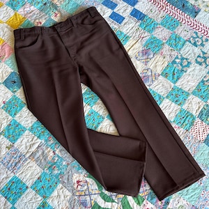 Puede incluir: Pantalones marrones con un diseño clásico de pierna recta. Los pantalones tienen cierre de botón y cremallera, trabillas para el cinturón y bolsillos delanteros. La tela tiene un ligero brillo y los pantalones están bien planchados. Se muestran sobre un edredón colorido.