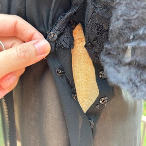 Peut inclure: Un chemisier noir transparent dot&eacute; d'une patte de boutonnage avant avec des boutons-pression noirs visibles. Le v&ecirc;tement pr&eacute;sente des d&eacute;tails en dentelle noire complexe le long de l'encolure et du corsage, en tissu l&eacute;ger semi-transparent.