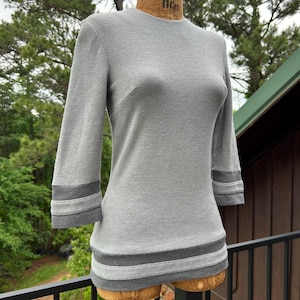 May include: A light gray, long-sleeve top with three-quarter sleeves. The top features a round neckline and is accented with darker gray bands at the cuffs and hem. The garment is displayed on a mannequin.