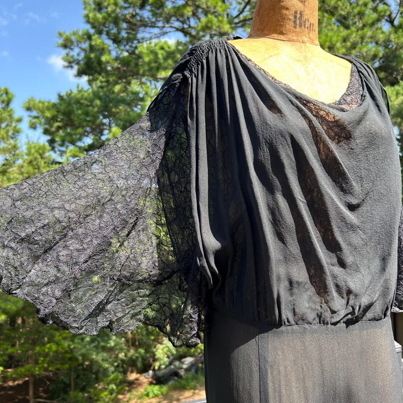 Peut inclure: Une robe noire dot&eacute;e de larges manches en dentelle transparente avec un motif floral d&eacute;licat. Le corsage est compos&eacute; d'un tissu l&eacute;ger et semi-transparent avec une encolure drap&eacute;e et une taille fronc&eacute;e, pr&eacute;sent&eacute;e sur un mannequin de couture beige.
