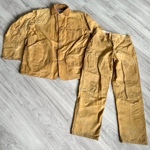 May include: A tan workwear jacket and trousers set. The jacket features a collared neckline, button fastenings, and multiple pockets. The trousers are straight-legged with a button and zip closure. The set is made of a durable, possibly waxed, fabric.