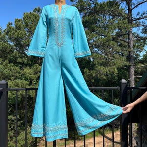 Op de afbeelding: Een turquoise jumpsuit met wijde pijpen en lange, uitlopende mouwen. Het vintage kledingstuk heeft ingewikkelde zilveren borduursels langs de halslijn, mouwen en broekzomen. De jumpsuit wordt buiten gepresenteerd.