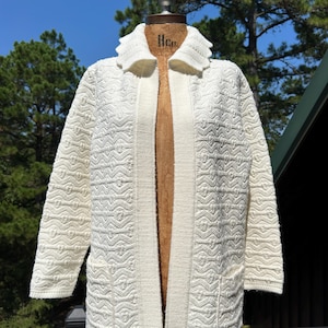 May include: Cream-colored, open-front cardigan sweater with a textured knit pattern. The long-sleeved sweater features a shawl collar and two front pockets. The garment is displayed on a vintage dress form.