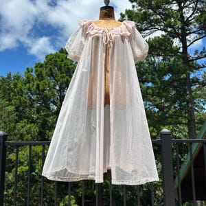 Pode incluir: Um roupão transparente em rosa pálido com mangas curtas bufantes, decote com folhos e fecho de laço, e acabamento em renda delicada na gola e na bainha. A peça leve abre-se na frente e é feita de um tecido fluido e semitransparente.