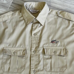 May include: A beige long-sleeve button-down shirt with two chest pockets and a fly fishing lure embroidered on the front. The shirt has the brand name "Walls" on the collar.