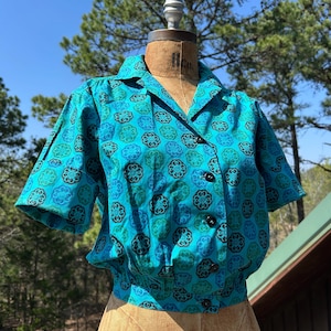 Puede incluir: Una blusa vintage turquesa con botones y un estampado floral en tonos azules y negros. La parte superior de manga corta presenta un cuello y botones negros. La blusa se exhibe en un maniquí.