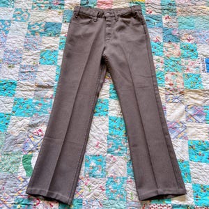 May include: Brown trousers with a button and zipper closure, belt loops, and front and back pockets. The pants are made of a solid-colored fabric and have a classic, straight-leg fit. The trousers are displayed on a colorful patchwork quilt.
