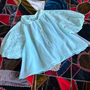 Puede incluir: Una bata vintage azul claro con mangas de encaje y cuello Peter Pan. La bata tiene tres botones blancos en la parte delantera y una tela transparente y fluida. Las mangas son de encaje blanco contrastante.