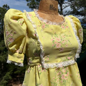 May include: A vintage yellow dress with a floral pattern of pink flowers and green leaves. The dress features short, puffed sleeves, a square neckline, and white lace trim. The dress is displayed on a mannequin.