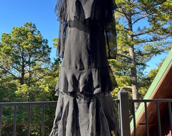 Peut inclure: Une robe noire vintage avec des volants superposés et des manches transparentes. La robe est présentée sur un mannequin en extérieur, avec une toile de fond d'arbres et un ciel bleu. La robe a une jupe à plusieurs niveaux et une silhouette fluide.