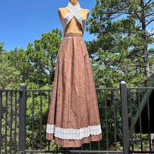 May include: A two-piece outfit consisting of a white crocheted halter top with a crisscross neckline and a floor-length brown skirt. The skirt features a subtle tonal floral pattern and a wide band of white lace trim along the bottom hemline.