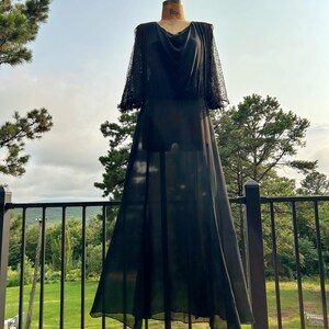 Peut inclure: Une robe noire longue jusqu'au sol avec une jupe et un corsage fluides et transparents, dot&eacute;s d'une encolure drap&eacute;e. Le v&ecirc;tement pr&eacute;sente des manches mi-longues en dentelle noire complexe, contrastant avec le tissu opaque du corps principal.