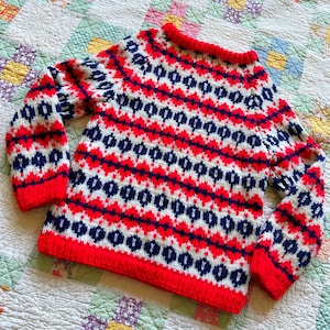 May include: A long-sleeve knit sweater featuring a repeating geometric pattern in red, white, and navy blue. The garment has a ribbed crew neckline, ribbed cuffs, and a ribbed hem, all in a solid red color, with a thick, textured knit construction.