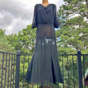 Peut inclure: Une robe noire longue jusqu'aux pieds avec des manches longues transparentes orn&eacute;es de dentelle, un d&eacute;collet&eacute; drap&eacute; et une jupe fluide. Le v&ecirc;tement est pr&eacute;sent&eacute; sur un mannequin, r&eacute;v&eacute;lant un tissu semi-transparent sur le corsage et la jupe.