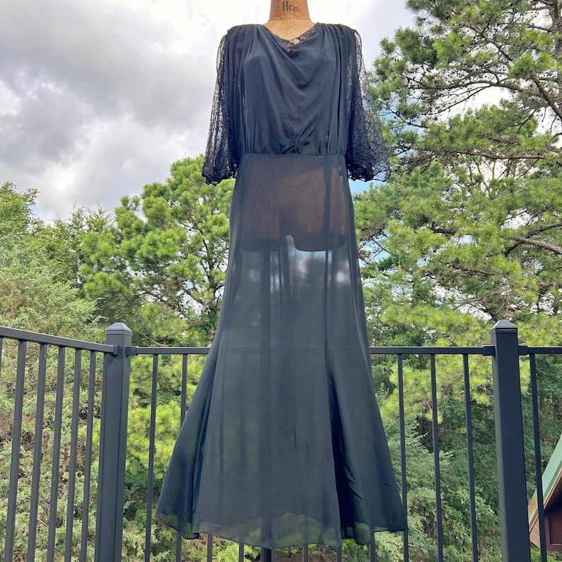 Peut inclure: Une robe noire longue jusqu'aux pieds avec des manches longues transparentes orn&eacute;es de dentelle, un d&eacute;collet&eacute; drap&eacute; et une jupe fluide. Le v&ecirc;tement est pr&eacute;sent&eacute; sur un mannequin, r&eacute;v&eacute;lant un tissu semi-transparent sur le corsage et la jupe.